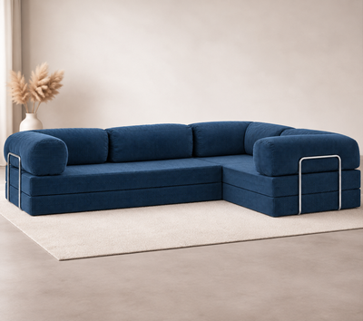 Confex Corner Blue Sofa sits across a cream rug against a pale living room wall, with deep blue cushions, curved armrests and silver-toned outer-arm details beside a vase of dried foliage.